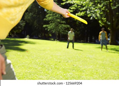 Cropped Hand In The Foreground About To Throw A Frisbee To Two People At The Background, With Trees In The Background