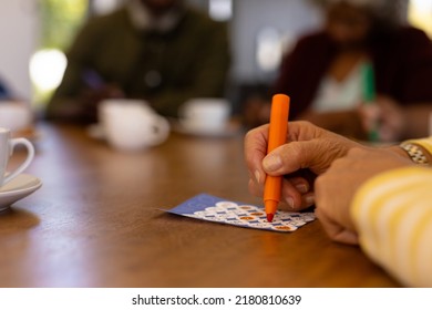 Cropped hands of biracial senior woman marking off numbers on bingo card over table in nursing home. Felt-tip pen, luck, leisure game, unaltered, support, assisted living, retirement concept.