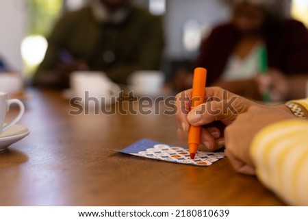 Cropped hands of biracial senior woman marking off numbers on bingo card over table in nursing home. Felt-tip pen, luck, leisure game, unaltered, support, assisted living, retirement concept.