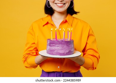 Cropped happy fun smiling young woman wearing casual clothes cap hat celebrating holding in hand purple sweet cake with candles isolated on plain yellow background. Birthday 8 14 holiday party concept