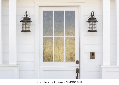Cropped horizontal shot of a white door to a classic, white, family home in daytime. The door has intricate, etched, glass detail. Also see is light fixtures, columns, and a door bell. 