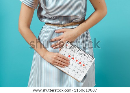 Cropped illness woman in blue dress holding periods calendar for checking menstruation days put hand on abdomen isolated on blue background. Medical, healthcare, gynecological concept. Copy space