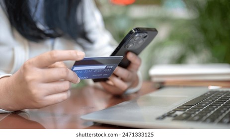 A cropped image of an Asian woman in casual clothes holding her smartphone and credit card at a table outdoors, using her mobile banking app to transfer her money. people and financial concepts - Powered by Shutterstock