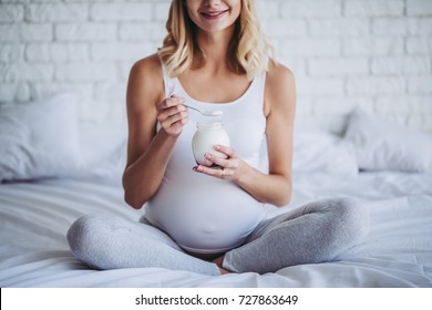 Cropped image of attractive pregnant woman is sitting in bed and eating yogurt. Healthy food concept. Last months of pregnancy.