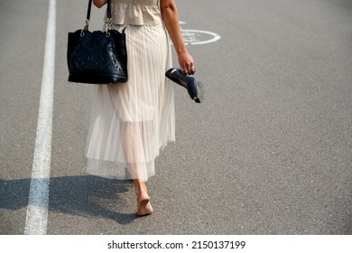 Cropped Image Of Bare Dirty Feet. The Woman Took Off Her Shoes, Holds The Shoes In Her Hands, And Walks Barefoot Along The Asphalt Road