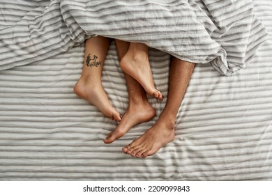 Cropped image of barefoot feet of man and woman with tattoo lying under blanket on bed. Top view of couple resting at home. Concept of domestic lifestyle. Idea of relationship. Bedroom. Daytime - Powered by Shutterstock