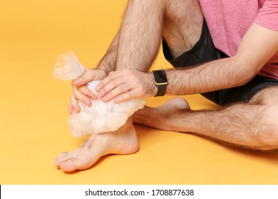 Cropped Image Of Bearded Fitness Sporty Guy 20s Sportsman In Shorts In Home Gym Isolated On Yellow Background. Workout Sport Motivation Concept. Get Injured Hold Ice Pack Cooling Down Ankle Foot Pain