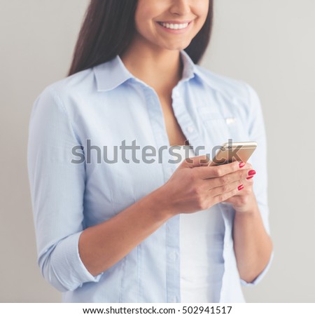 Cropped image of beautiful business lady in smart casual wear using a smartphone and smiling, on a gray background