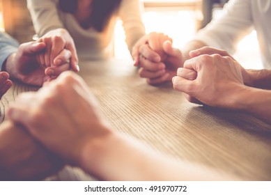 Обрезанное изображение красивой бизнес-команды, держащейся за руки и молящейся, сидя в офисе