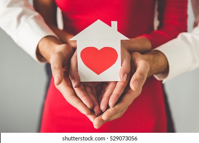 Cropped Image Of Beautiful Elegant Couple Holding A Paper House With Heart, On Gray Background