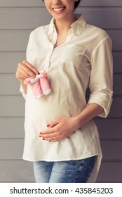 Cropped image of beautiful pregnant woman holding baby shoes, keeping one hand on a belly, looking at camera and smiling, standing against gray wall - Shutterstock ID 435845203