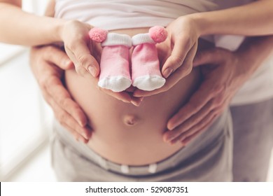 Cropped image of beautiful pregnant woman and her handsome husband holding baby shoes and hugging the tummy
