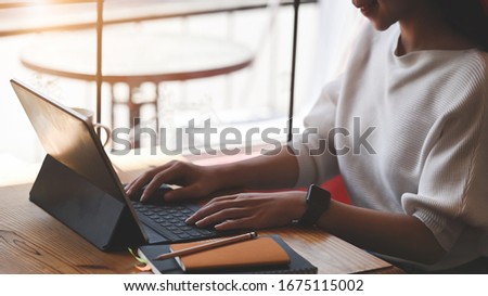 Cropped image of beautiful woman typing on computer tablet with keyboard case while sitting at the orderly wooden table over living room as background.