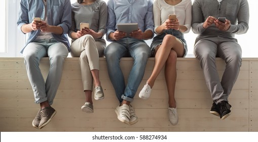 Cropped Image Of Beautiful Young People In Casual Clothes Using Gadgets While Sitting Together On The Window Sill