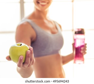 Cropped image of beautiful young sports lady smiling, holding an apple and a bottle of water