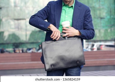 Cropped image businessman in casual wear with cup of coffee to go puts smartphone into trendy leather briefcase against modern office building background. - Powered by Shutterstock