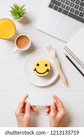 Cropped Image Of Businesswoman Holding Empty Visit Card At Table With Orange Juice, Coffee Cup And Cake With Symbol Of Smile In Office