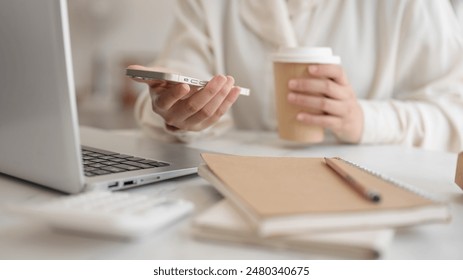 A cropped image of a businesswoman working remotely at a cafe, using her smartphone, a laptop and a coffee cup are on the table. - Powered by Shutterstock