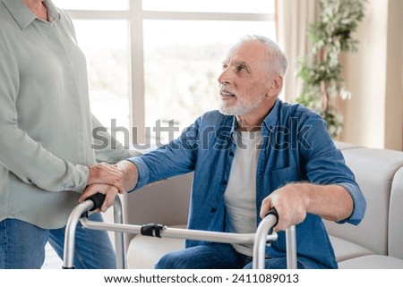 Cropped image of caregiver caretaker wife helping aiding supporting old elderly senior incapacitated handicapped disabled husband patient with walking frame at home hospice clinic