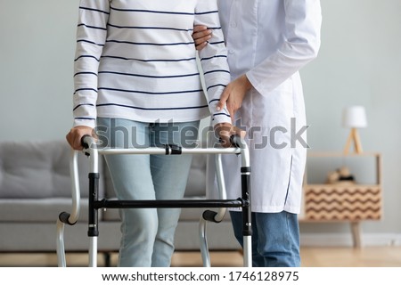 Cropped image caregiver helping to disabled aged female patient walk with walker physical therapist teach her, supporting during exercise therapy. Nursing at home, aftercare and physiotherapy concept