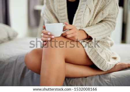 Cropped image of caucasian woman in sweater holding cup of coffee while sitting on bed in bedroom.