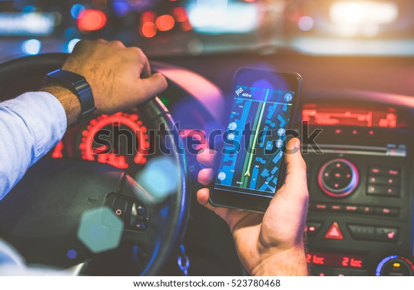 Cropped\
image. Closeup of smartphone in hand of young man sitting behind\
the wheel of modern car. Guy uses digital gadget. Man paves way to\
browser. On screen of smartphone map of\
area.