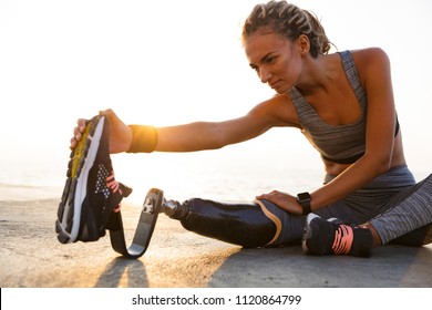 Cropped image of concentrated disabled athlete woman with prosthetic leg doing stretching exercises while sitting at the beach - Powered by Shutterstock