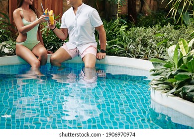 Cropped image of couple sitting by hotel swimming pool among lush plants and drinking delicious fruit cocktails