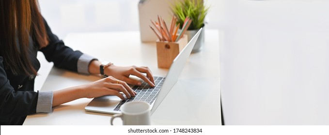 Cropped Image Of Creative Woman Hands Typing On A Computer Laptop That Putting On A White Working Desk Surrounded By A Coffee Cup, Pencil Holder, Potted Plant, And File Folder.