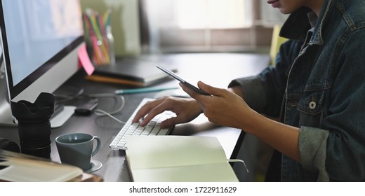 Cropped Image Of Designer Man Holding/looking A Smartphone In Hand While Typing On Keyboard And Sitting In Front A White Blank Screen Computer Monitor Over Cluttered Working Desk As Background.