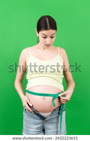 Cropped image of expecting mother in unzipped jeans measuring her pregnant belly with a tape measure at colorful background. Centimeter measurement.