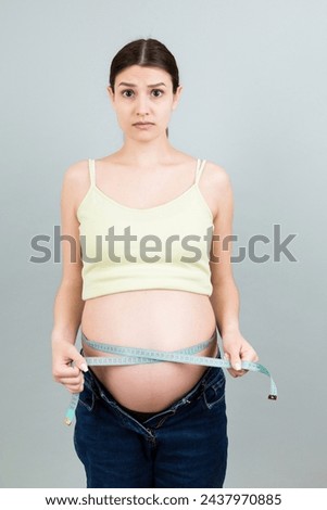 Cropped image of expecting mother in unzipped jeans measuring her pregnant belly with a tape measure at colorful background. Centimeter measurement.