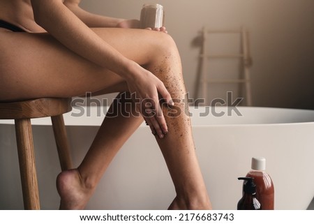 Cropped image female hand applying peeling organic coffee scrub on shin, massaging legs over ceramic bathtub in luxury bathroom. Beauty and spa procedures. Body and skin care
