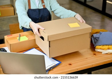 Cropped Image, Female Seller Holding A Big Cardboard Box Or Brown Parcel Box Over Her Desk, Packing The Customer's Order. Fashion Shop Owner.
