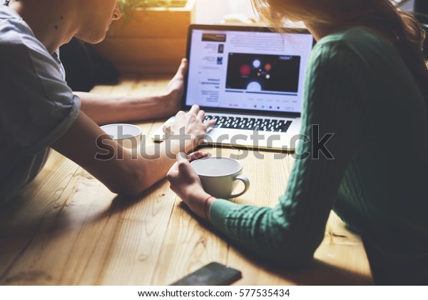 Cropped image of group of young students are\
watching movie on laptop computer, while are sitting at wooden\
table in loft. Group of friends using net-book during meet in\
modern coffee shop\
interior