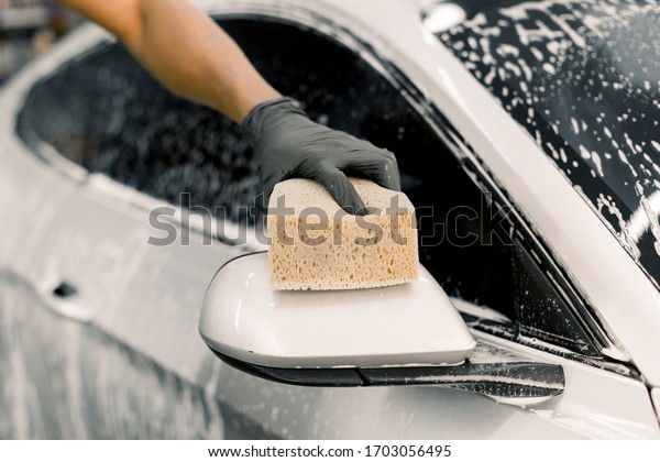 Cropped image of hand of car wash worker washing\
a soapy mirror of white luxury car, holding sponge. Focus on the\
hand in black glove, holding sponge and washing car mirror.\
Cleaning, car wash\
concept