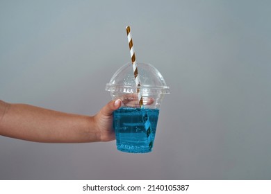 Cropped Image Of Little Girl Hand Holding Plastic Cup With Tubule And Soda Drink. Blue Fresh Sweet Drink. Childhood Concept. Isolated On Grey Background. Studio Shoot. Copy Space