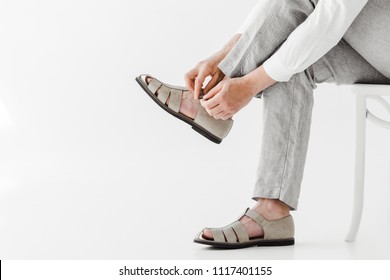 Cropped Image Of Male Model In Linen Trousers Sitting On Chair And Putting On Sandals Isolated On Grey Background 