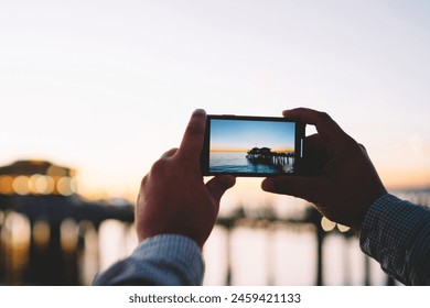 Cropped image of man making picture of environment and sea at evening having good app on mobile phone, male taking image of ocean in twilight via modern smartphone and camera for photo in dark