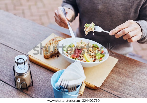 Cropped image\
of a man on his lunch break eating a fresh and healthy salad with\
chicken, avocado, sundried tomatoes and fresh sliced baguette on\
the side while sitting at a wooden\
table