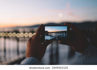 Cropped image of man taking photo in evening dusk of nature environment using modern smartphone camera, back view of hipster guy holding mobile phone making picture of sea water via app in twilight - Powered by Shutterstock