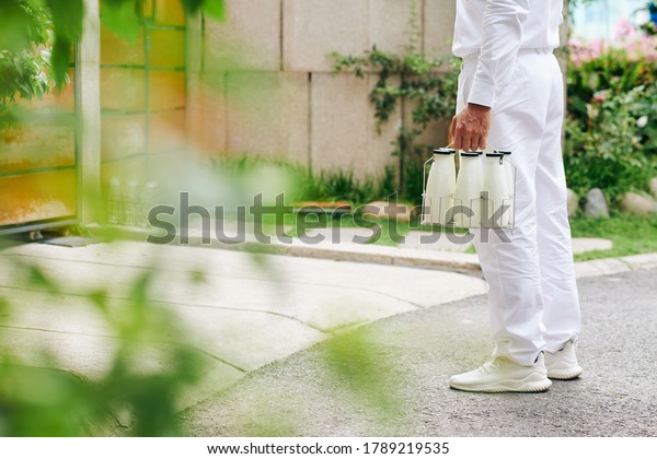 Cropped image of milkman\
in white uniform standing at house entrance with glass bottles of\
milk or yogurt