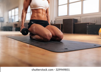 Cropped image of muscular woman exercising with dumbbells while sitting on fitness mat in gym. Focus on abs.
