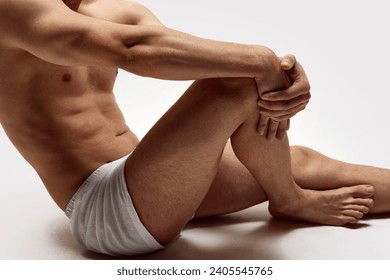 Cropped image of muscularly young shirtless man with relief body sitting in underwear, boxers isolated over white studio background. Concept of male beauty, body care, fitness, sport, health - Powered by Shutterstock