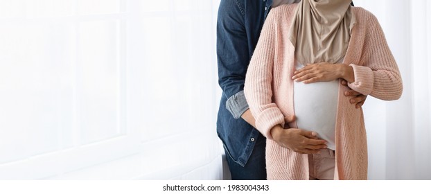 Cropped image of muslim couple expecting baby hugging near window at home, loving husband embracing pregnant wife's belly, islamic spouses cuddling and enjoying future parenting, panorama, copy space - Shutterstock ID 1983004343