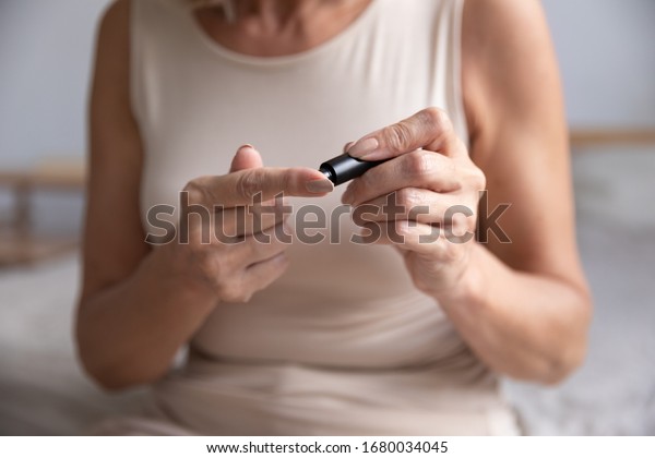 Cropped image older woman using glucose meter\
alone at home. Unhealthy middle aged retired lady testing blood\
sugar ranges in morning, putting lancet pen on finger. Diabetes,\
healthcare concept.