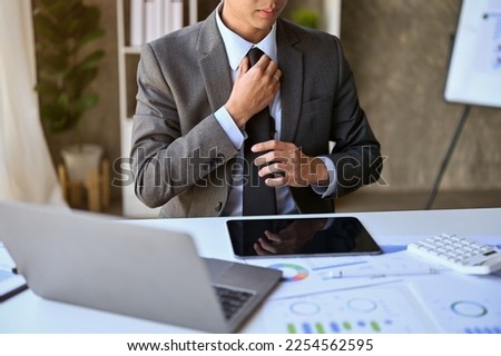 cropped image of a professional and successful millennial Asian businessman or male CEO in formal business suit, adjusting his tie at his desk.