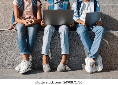 Cropped image of school kids preteen boys and girls elementary middle pupils sitting on the stairs and using digital devices wireless gadgets laptop tablet smartphone outdoors at the street - Powered by Shutterstock