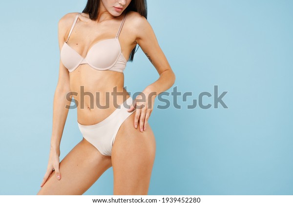 Cropped image of sexy attractive young brunette\
woman 20s in beige underwear showing fit body standing posing hold\
hand on hips legs isolated on pastel blue colour wall background\
studio portrait