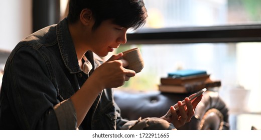 Cropped image of smart man drinking a hot coffee and holding/using a white blank screen smartphone while sitting and relaxing at the black leather sofa over comfortable sitting room as background. - Powered by Shutterstock